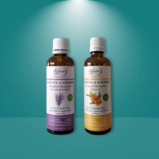 Two amber glass bottles of Ayluna Naturals Face & Body Oil side by side on a teal gradient background with a spotlight effect at the top. The left bottle has a white cap and a purple label, and the right bottle has a white cap and a yellow-orange label.