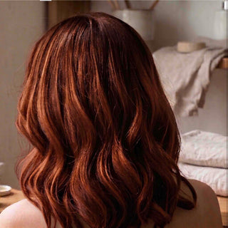 A close-up back view of a person with shoulder-length wavy hair in a warm coppery auburn shade, set against a softly blurred indoor background.