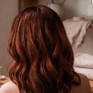 Medium-length wavy hair in a warm auburn brown shade, shown from the back on a person in a softly lit indoor setting.