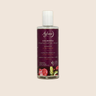 A bottle of Ayluna Shampoo for damaged hair, featuring a deep burgundy label with white text and illustrations of pomegranate and green leaves, topped with a white cap, set against a solid pale beige background.