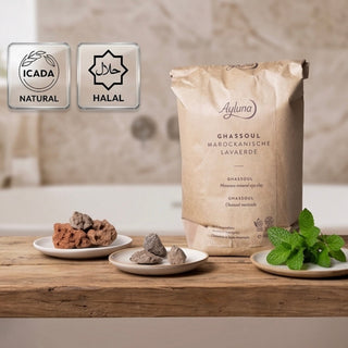 A bag of Ayluna Ghassoul Moroccan lava clay stands on a wooden table beside small plates of clay pieces and mint leaves. Two certification-style badges at the top left read ICADA NATURAL and HALAL.
