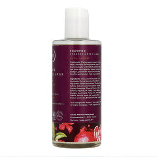 A bottle of natural shampoo for dry, frizzy, and damaged hair, featuring a white cap and a dark purple label with German text and illustrations of red cherries and green lime slices at the bottom.
