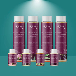 A set of Ayluna bath products arranged on a teal gradient background with a spotlight effect. The collection includes four large bottles of bath salts, two medium bottles, and two small bottles, all featuring deep purple labels with white caps and botanical illustrations.